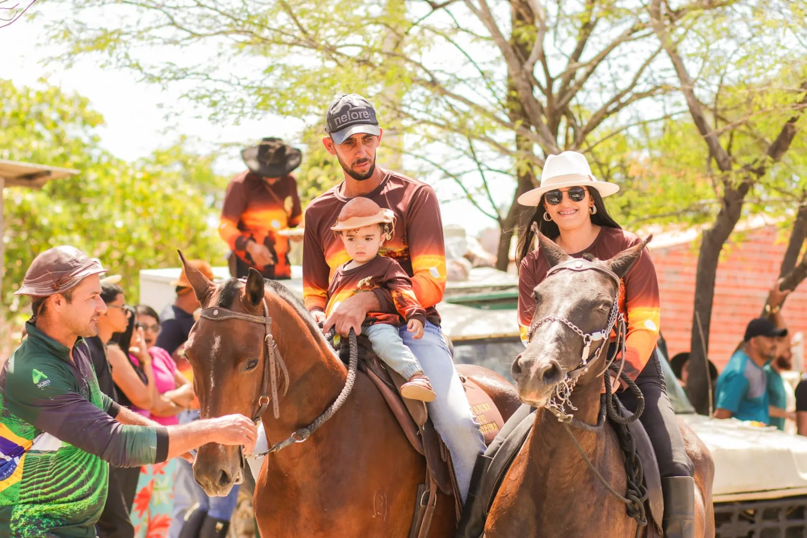 Pai com o filho pequeno no colo e mulher de chapéu branco montados a cavalo num evento ao ar livre