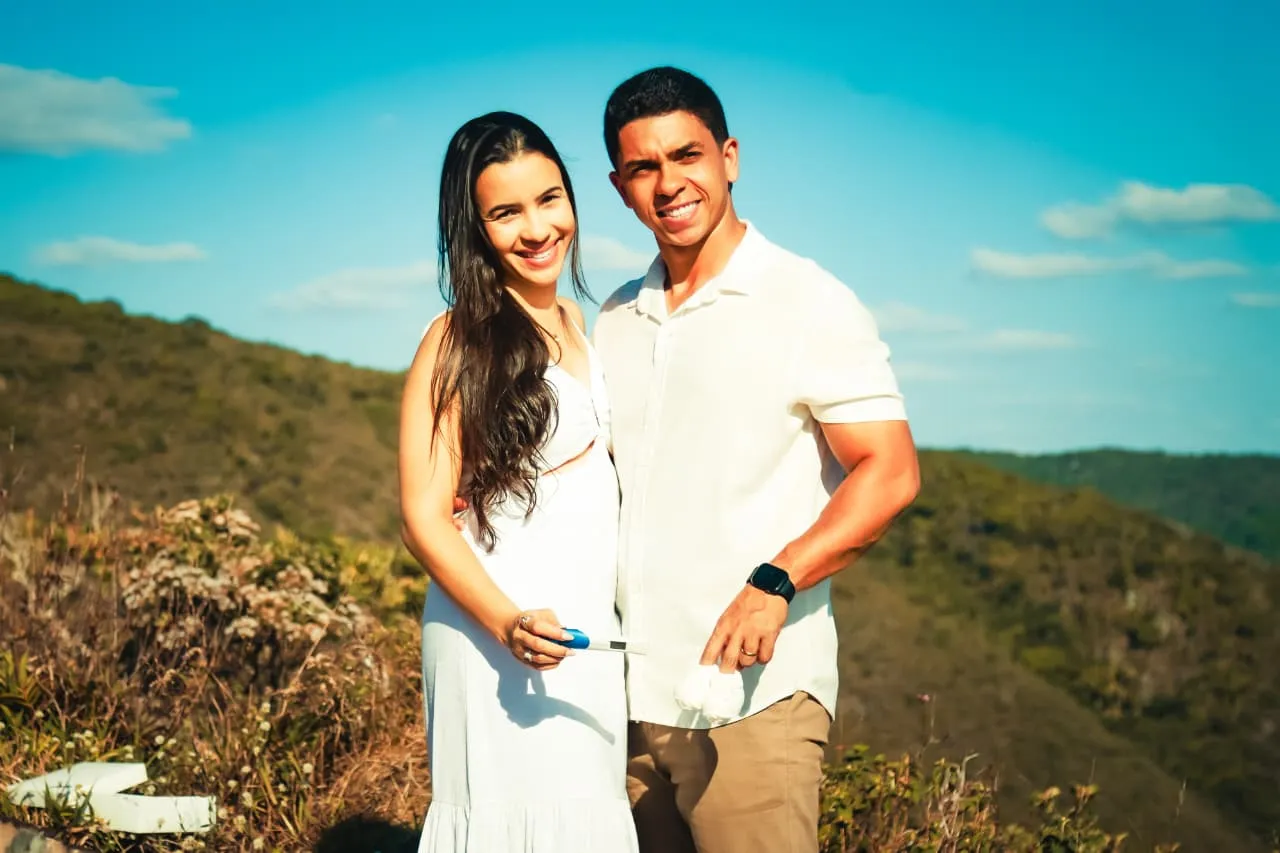Casal sorridente vestido de branco num ensaio ao ar livre, com montanhas e céu azul ao fundo